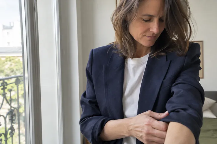 A woman in her forties in a navy unlined blazer over a white tee adjusts her sleeve cuff beside a bright window.